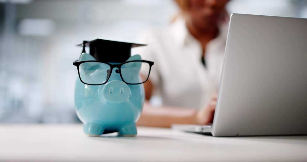 A close-up photo of a light blue glass piggy bank with a tiny black graduation hat and black-rimmed glasses sitting on a desk with a woman working on a laptop blurred in the background.