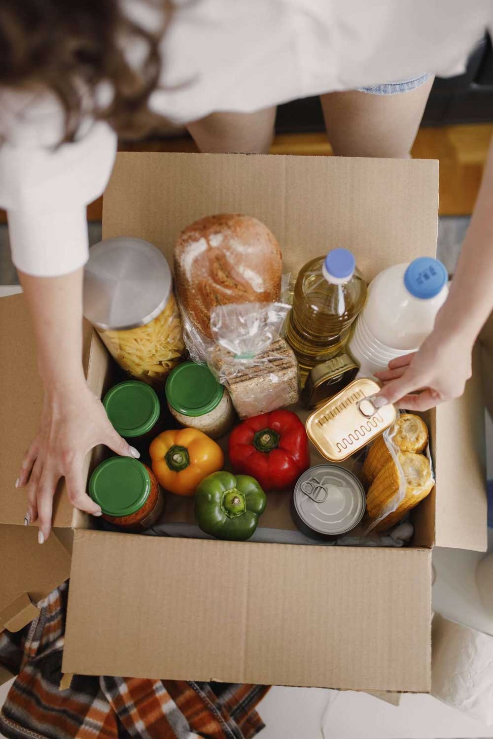 An overhead shot of a box of healthy food, including peppers, canned tuna, pasta, whole wheat bread, and milk.