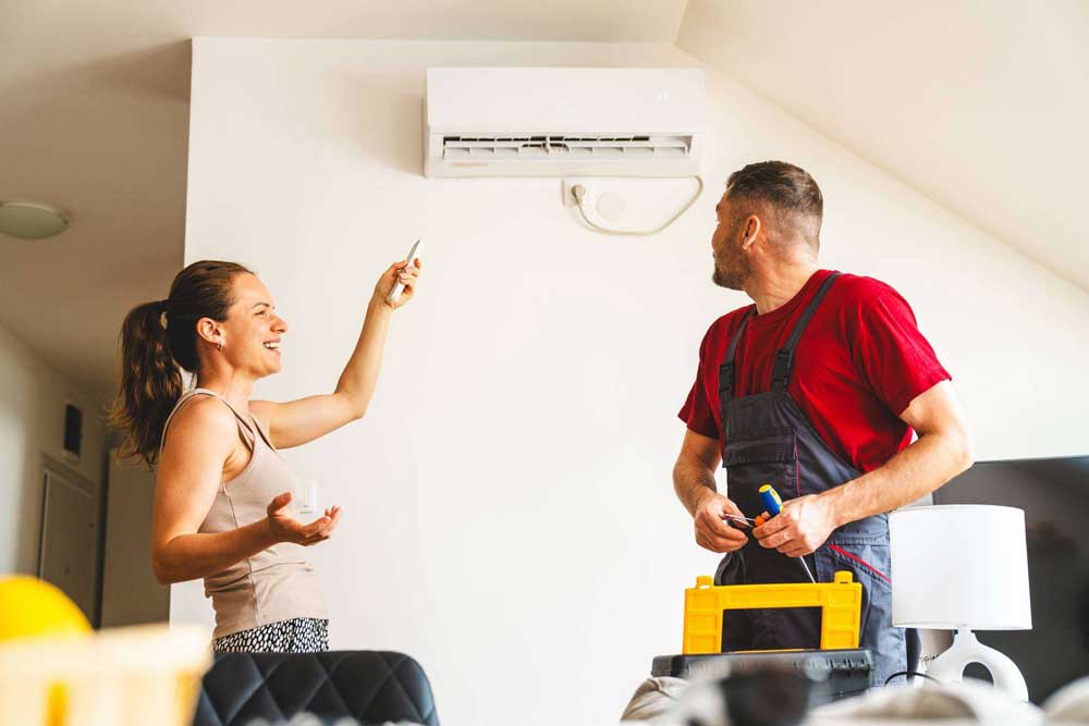 A happy, smiling woman checks the airflow coming from the newly reparied air conditioner in her living room while a male HVAC technician finishes putting his tools away.