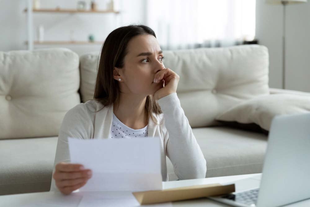 A young woman sits on the ground in front of her couch, opened mail and a laptop on the coffee table in front of her. She looks off into the distance, looking worried.