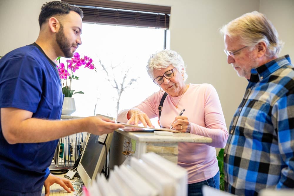 An older couple stand at the front desk of a medical office, signing off on a paper handed to them by a young man in scrubs.
