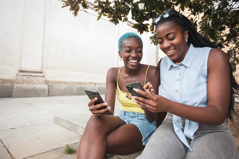 Two young Black women sit side by side on a cement bench outside a building, holding their phones together and smiling.