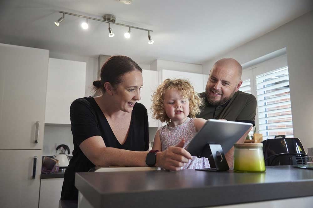 A young mom and dad stand in their kitchen with their toddler between them, smiling as they look at a tablet.