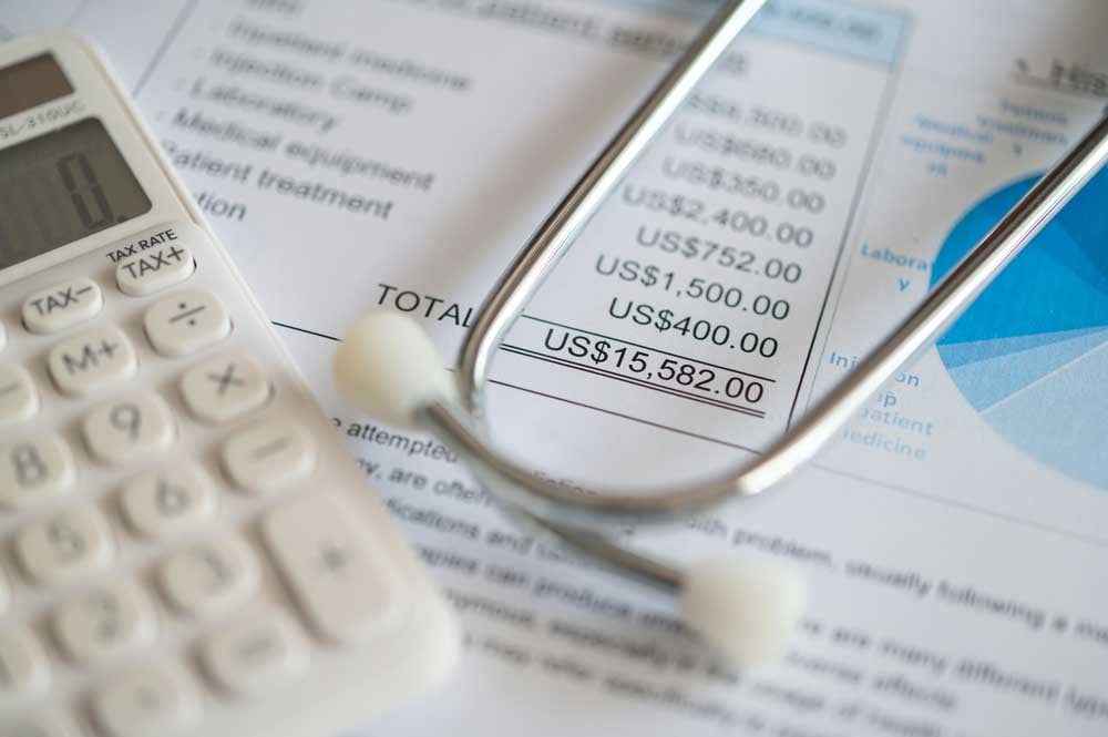 A close-up photo of an itemized medical bill totaling over $15,000. Slightly blurred in the foreground is part of a calculator and the ear tubes of a stethoscope.