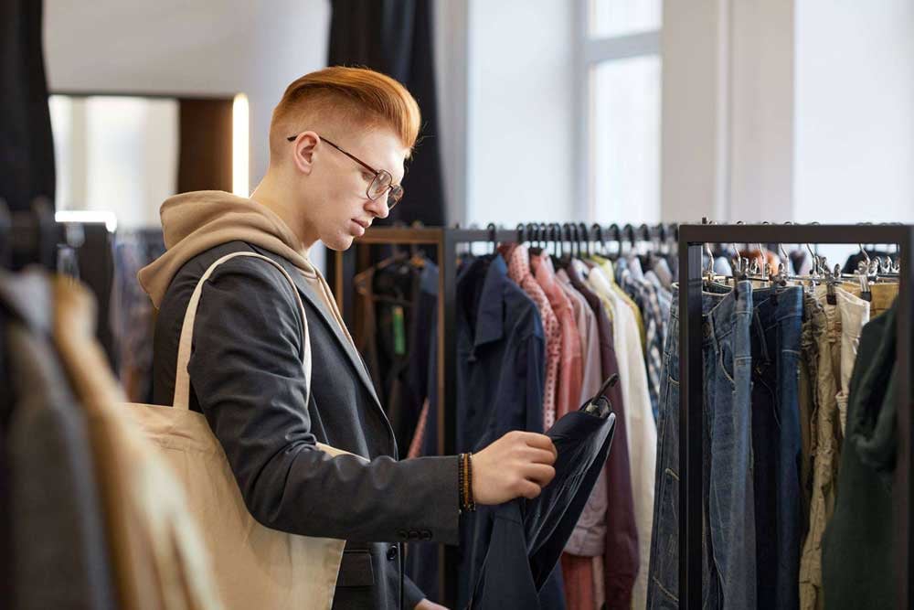 A photo of a young man in profile, looking at racks of jeans and khakis while shopping sustainably at a thrift store.