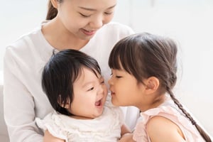 A photo of a young mom with a smiling infant in her lap and her young daughter leaning in to kiss her baby sister on the cheek. 