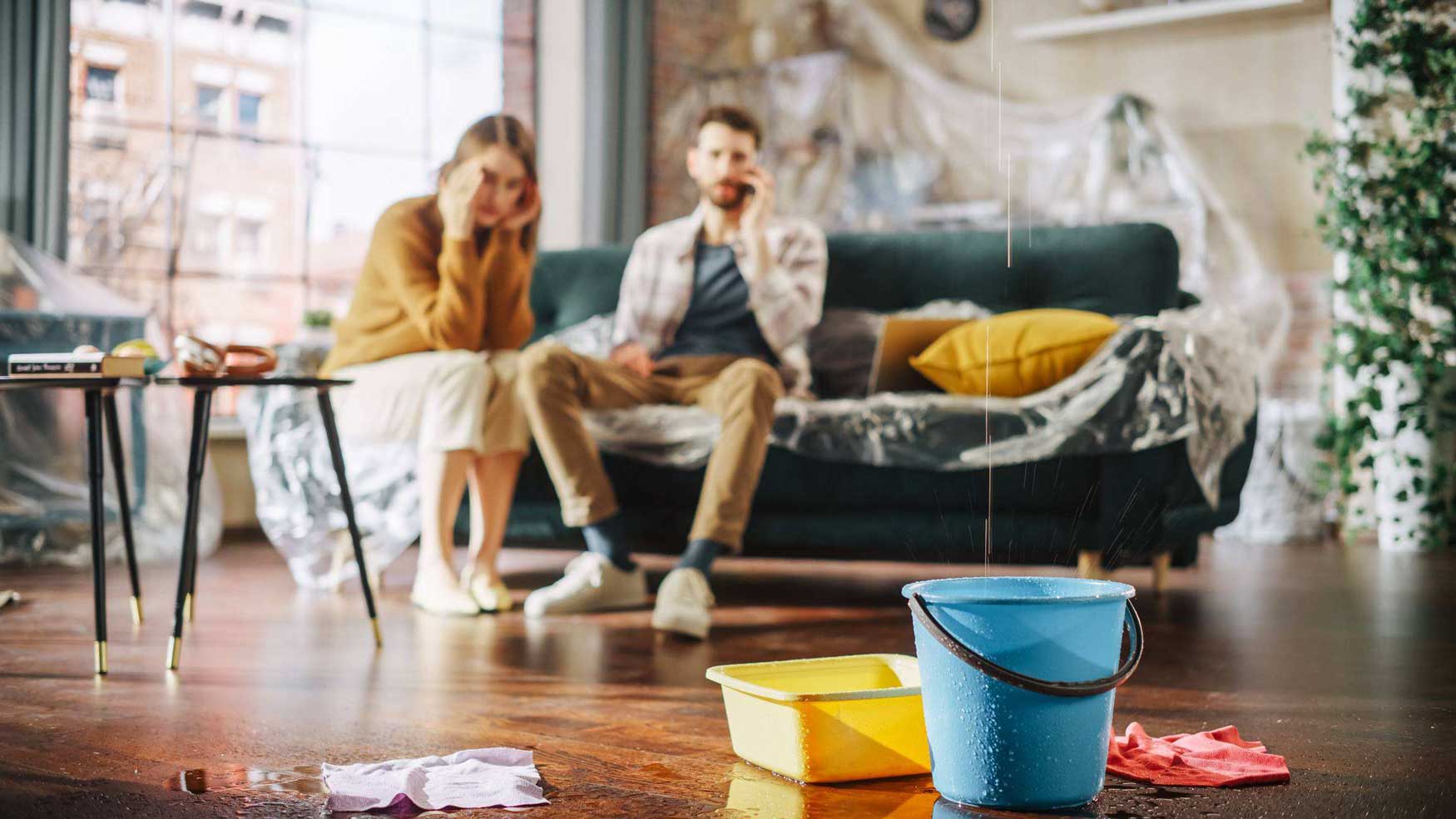 In the foreground, a plastic bucket sits under dripping water from a roof leak. In the blurred background, a couple sits looking at the mess, the man with a phone to his ear, calling a specialist. 