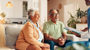 A senior couple sit on a couch, holding hands and smiling up at a young woman with a tablet, consulting with them on assistance programs.  