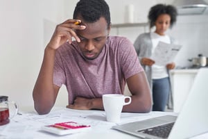A young man sits at a kitchen table, looking stressed. The table is scattered with bills, coffee mugs, and a calculator. His wife stands in the background, looking at another piece of paper.