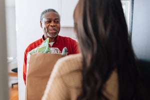 A woman with long dark hair stands in a doorway, handing a bag of groceries to a smiling elderly man.