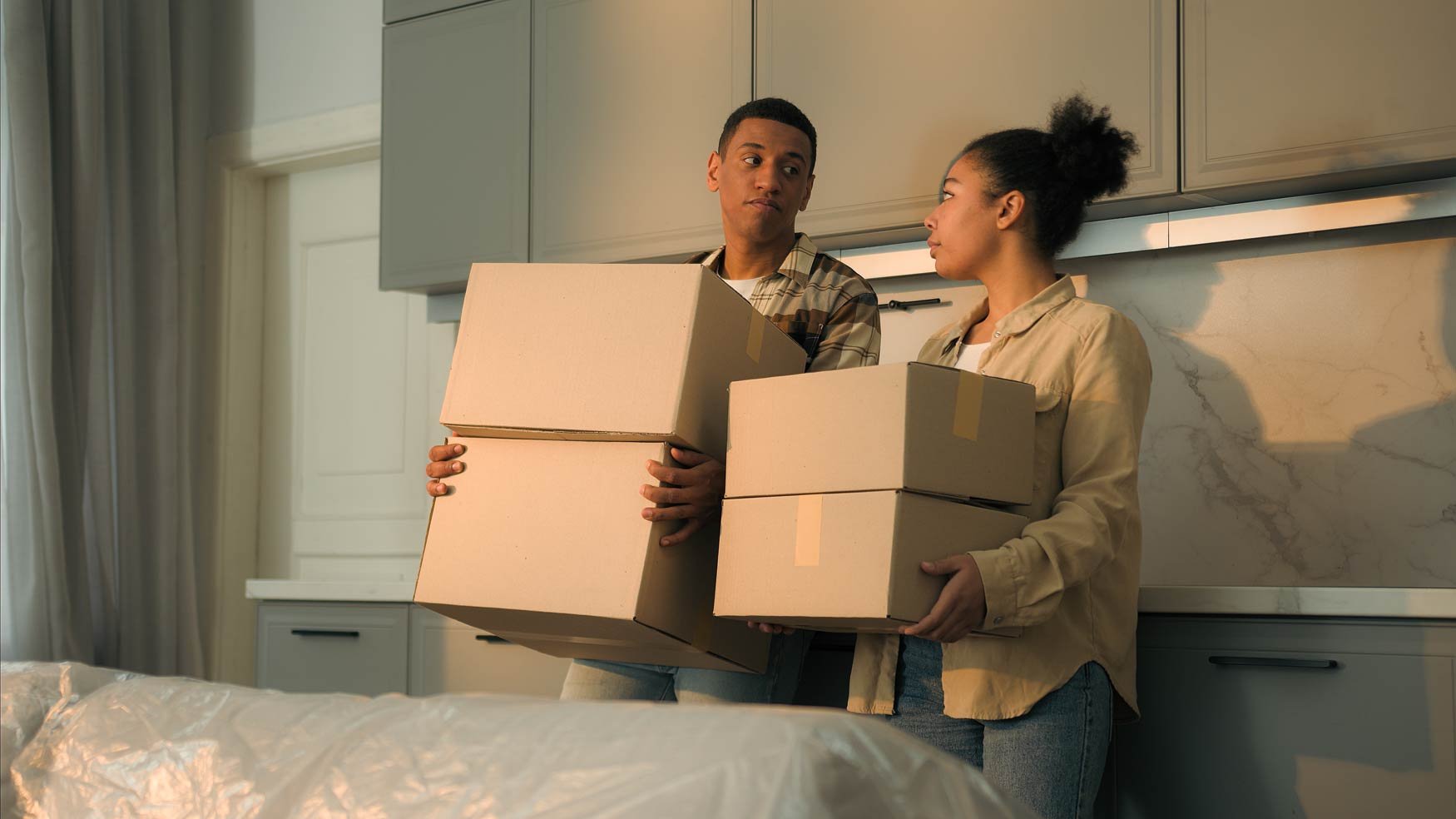 A young couple stands in a kitchen holding moving boxes, looking at each other with concern. 