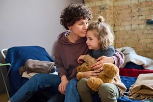 A mother with brown curly hair sits on a low cot, holding her young daughter who is cuddling a teddy bear.
