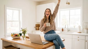 A smiling woman sits at her kitchen table in front of her computer with a coffee. Her are arms up in the air in celebration. 