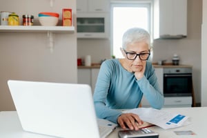 An older woman with short white hair sits in her kitchen, working a calculator with her laptop in front of her.