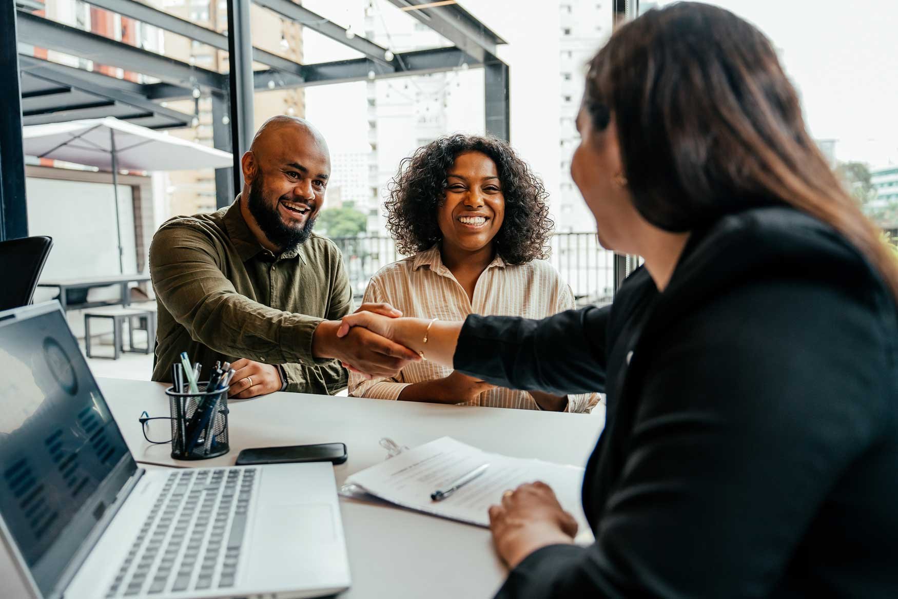 A smiling couple shakes hands with a woman in a suit who is sitting in front of a laptop.