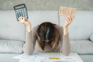 A young woman sits on the floor with bills all over the coffee table and her head down, holding a calculator in one hand and a sign that says “Help” in the other.
