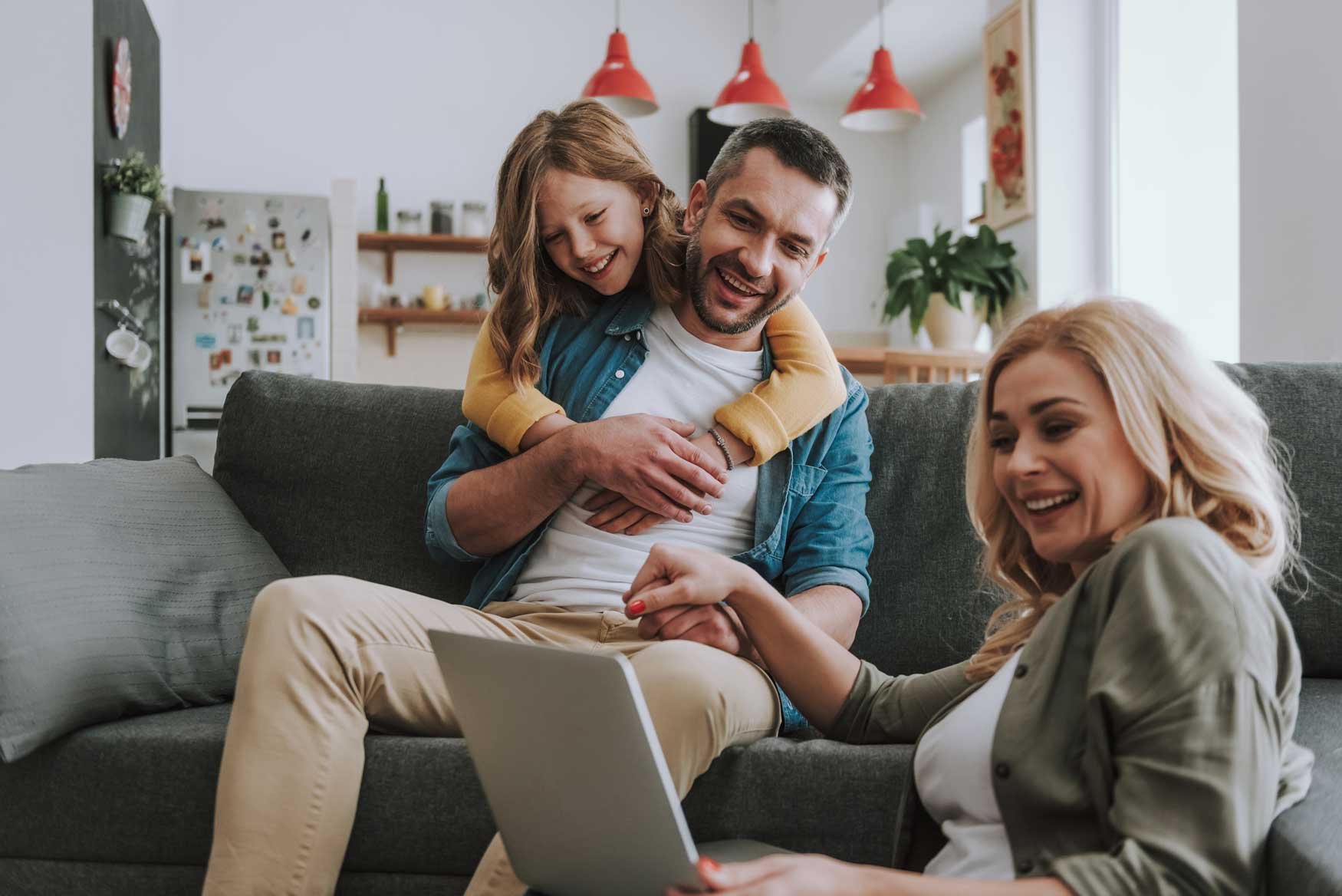 A smiling family sits in their living room, the mother on the ground with a laptop, the father on the couch with their daughter draped over his shoulders. 
