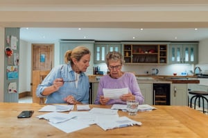 An elder woman sits at a kitchen table with her middle-aged daughter looking over medical bills and financial records.
