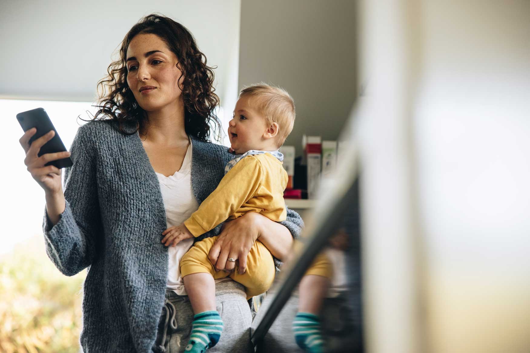 A mother stands in the doorway of a pantry with a baby on her hip, checking her phone with a small smile on her face. 
