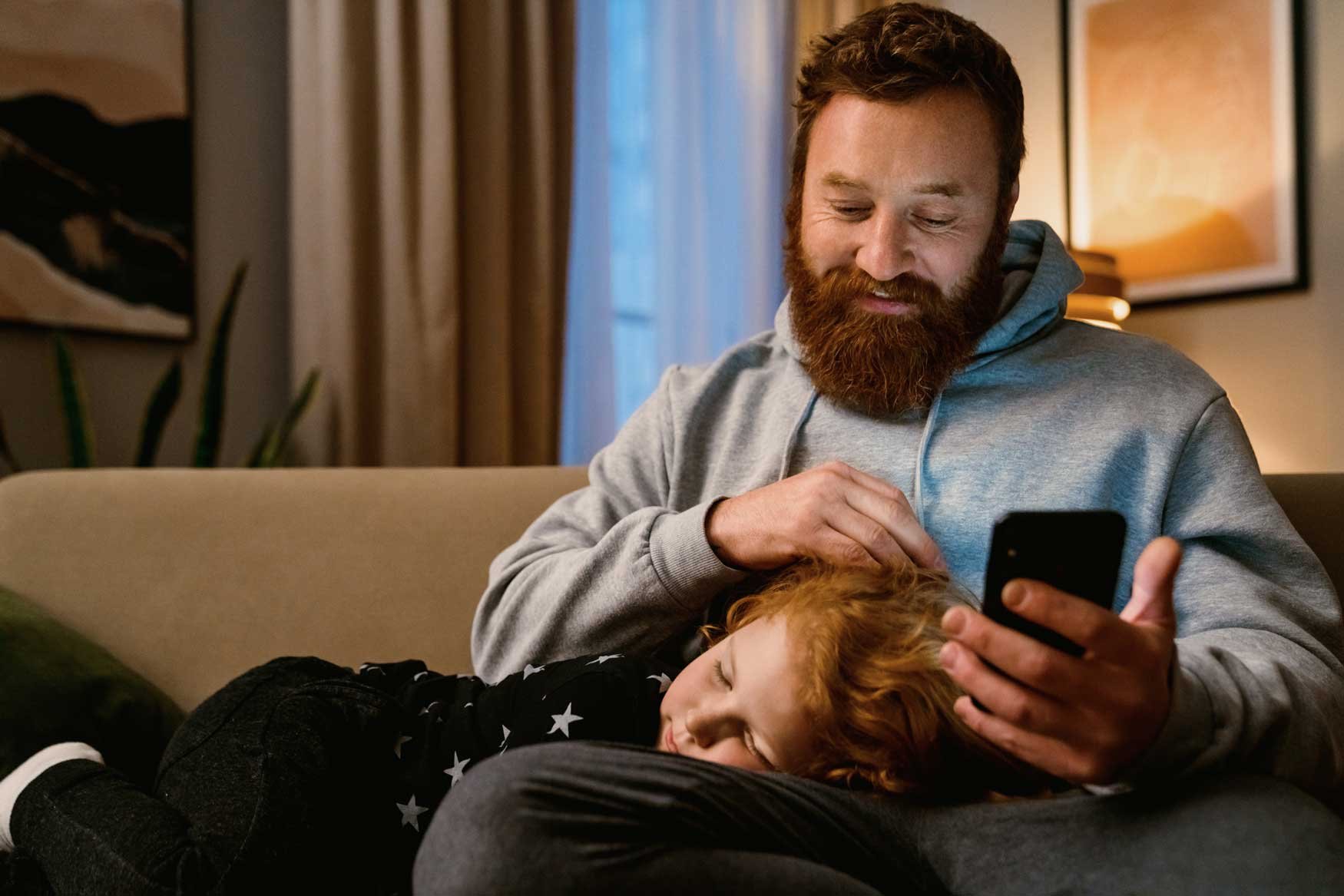 A father sits on his couch, his small child asleep with their head in his lap. 