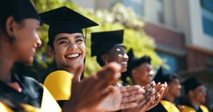 A photo of a line of college graduates in caps and gowns smiling and clapping with focus on a young woman in the center turned toward the camera to smile at her friend. 