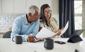 A man and woman in their 60s sit side by side at their kitchen table, reviewing their taxes and financial documents.
