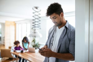 A man stands in a kitchen looking down at his smartphone with his family blurred in the background. 