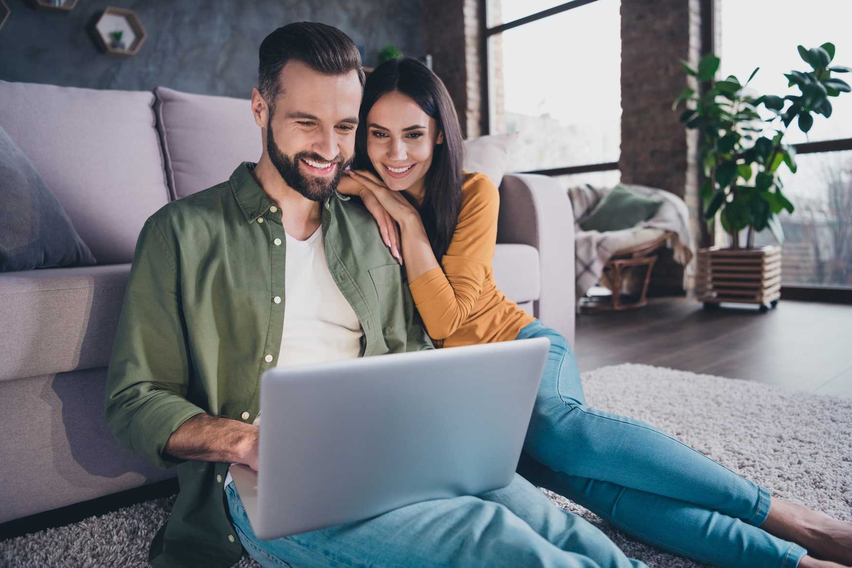 A smiling couple sits on their living room floor in front of their couch, both looking at the laptop in the man’s lap.
