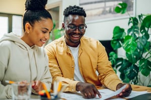 Two young people sit together, both smiling as they look over paperwork on the table in front of them. 
