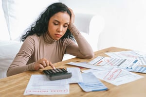 A young brunette woman sits at a table, typing on a calculator, overdue bills strewn in front of her.