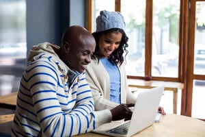 Two young people, close to college age, stand together in a cafe, smiling and looking at the laptop on the table in front of them. 