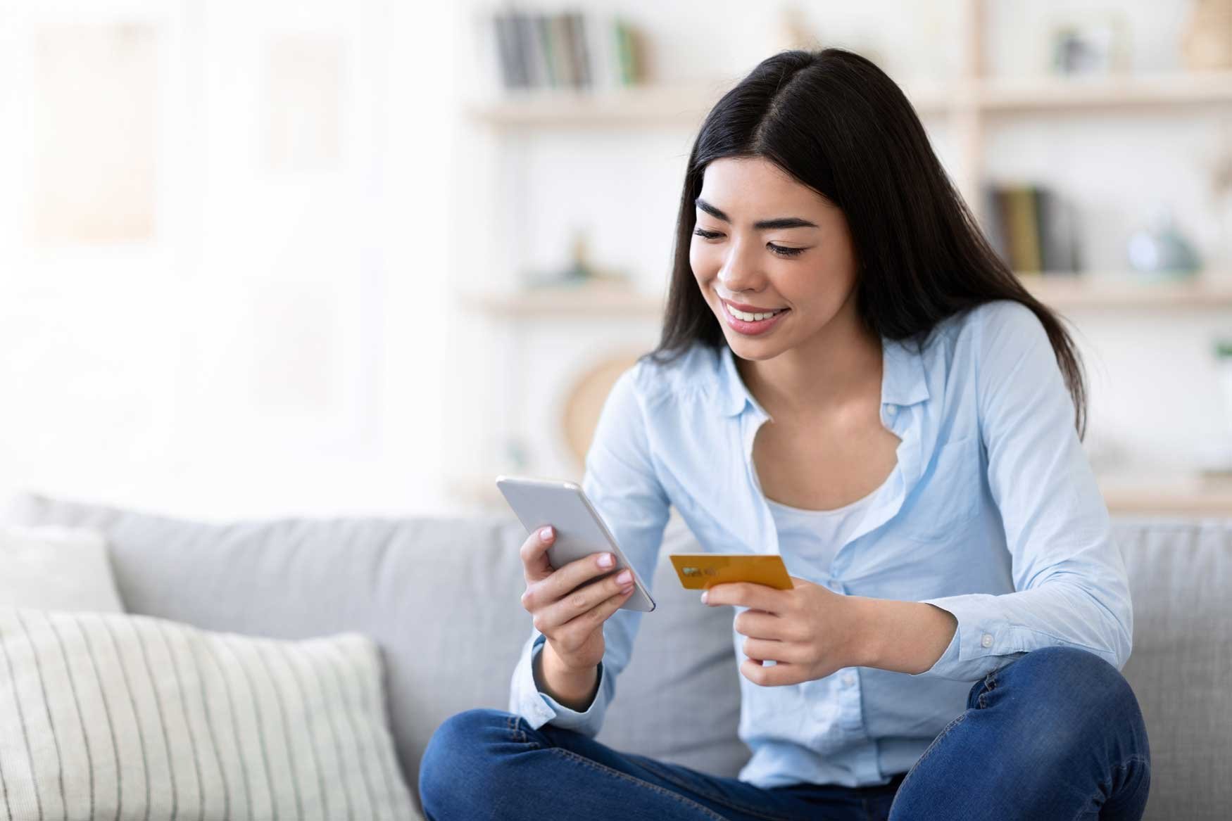 A young woman sits on her couch, smiling, her smartphone in one hand and a credit card in the other. 