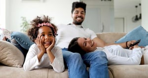A smiling family sits on the couch, the young daughter on her stomach with her chin propped up on her hands with her parents in the background.