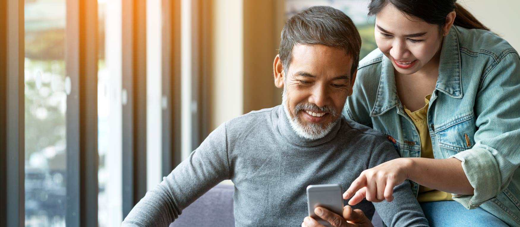 A middle-aged man smiles and looks at his smartphone as his teen daughter leans over his shoulder to point something out to him. 