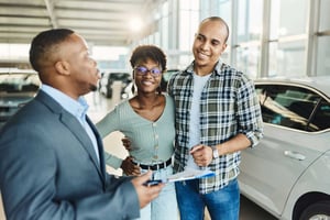 Young, smiling African American couple talks with a car salesperson in a dealership showroom about buying a new car. 