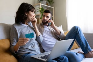 A couple sits together on the couch, the wife in the foreground holding a laptop and a credit card, the man next to her on the phone. They both look shocked.