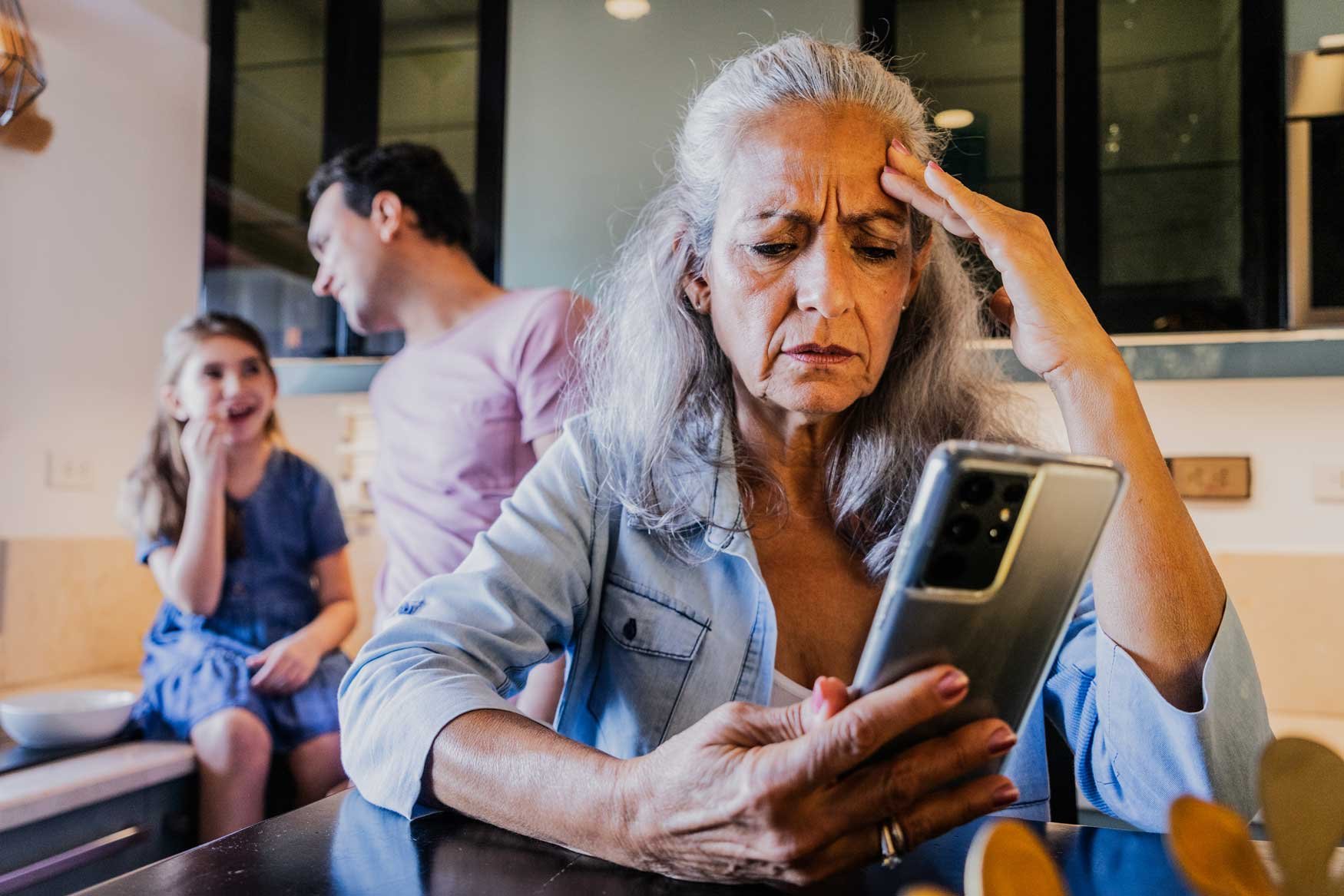 An elderly woman in the foreground looks at her phone looking very stressed while her son and grandchild stand in the background. 