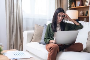 A young woman sits on a white couch with her laptop in her lap, looking stressed. 