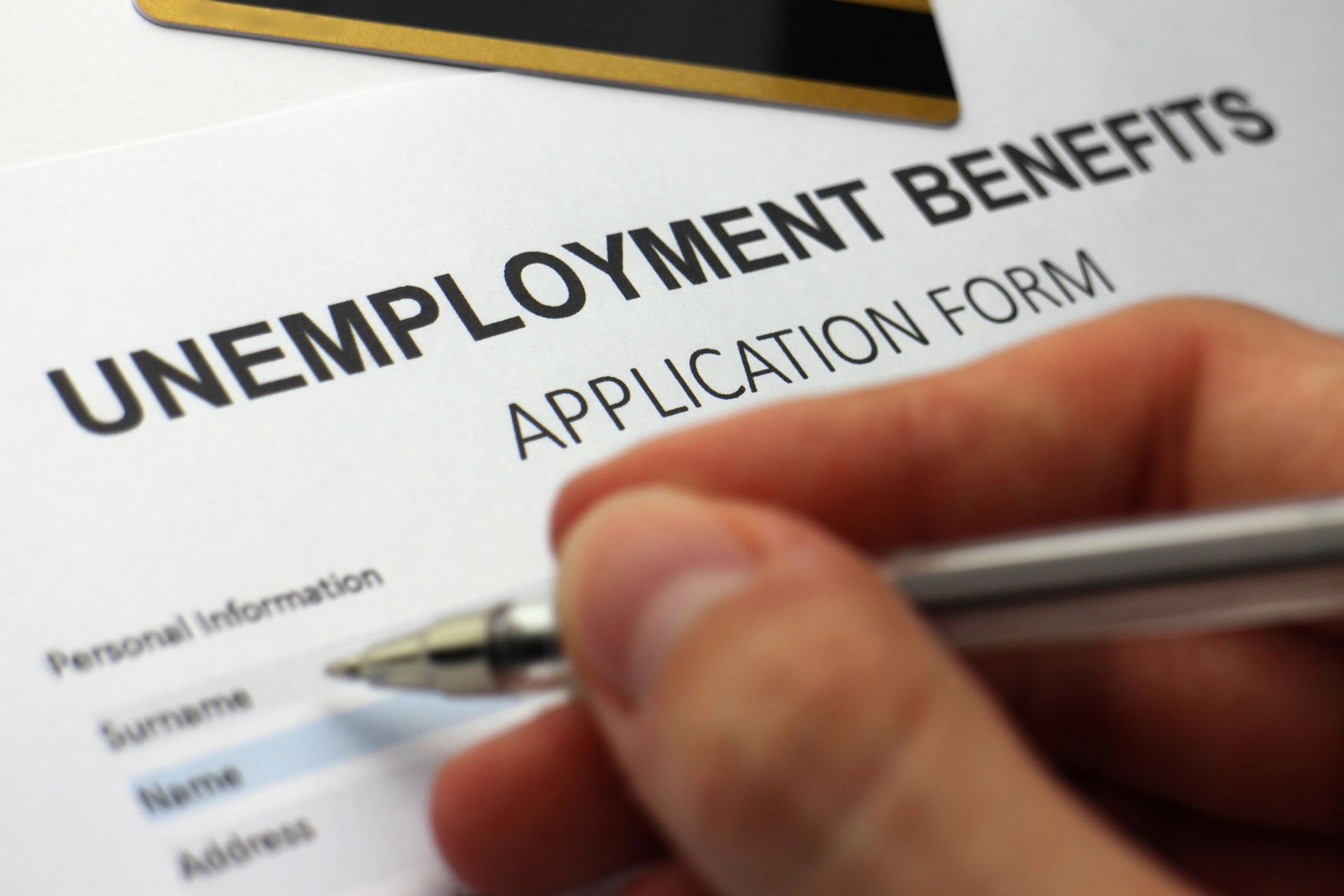 A close-up photo of a hand filling out a paper form with a pen. At the top of the form are the words: Unemployment Benefits Application Form.