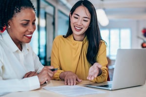 A young woman in a white button down sits at a table with a young woman in a yellow shirt who is walking her through the paperwork on the table in front of them. 
