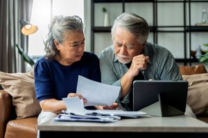 An older couple sits on their couch looking down with concern at the bills and opened envelopes on the coffee table in front of them. 