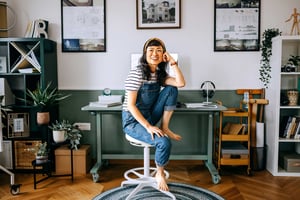 A smiling young Asian woman sits barefoot in her home office with her desk and computer behind her.